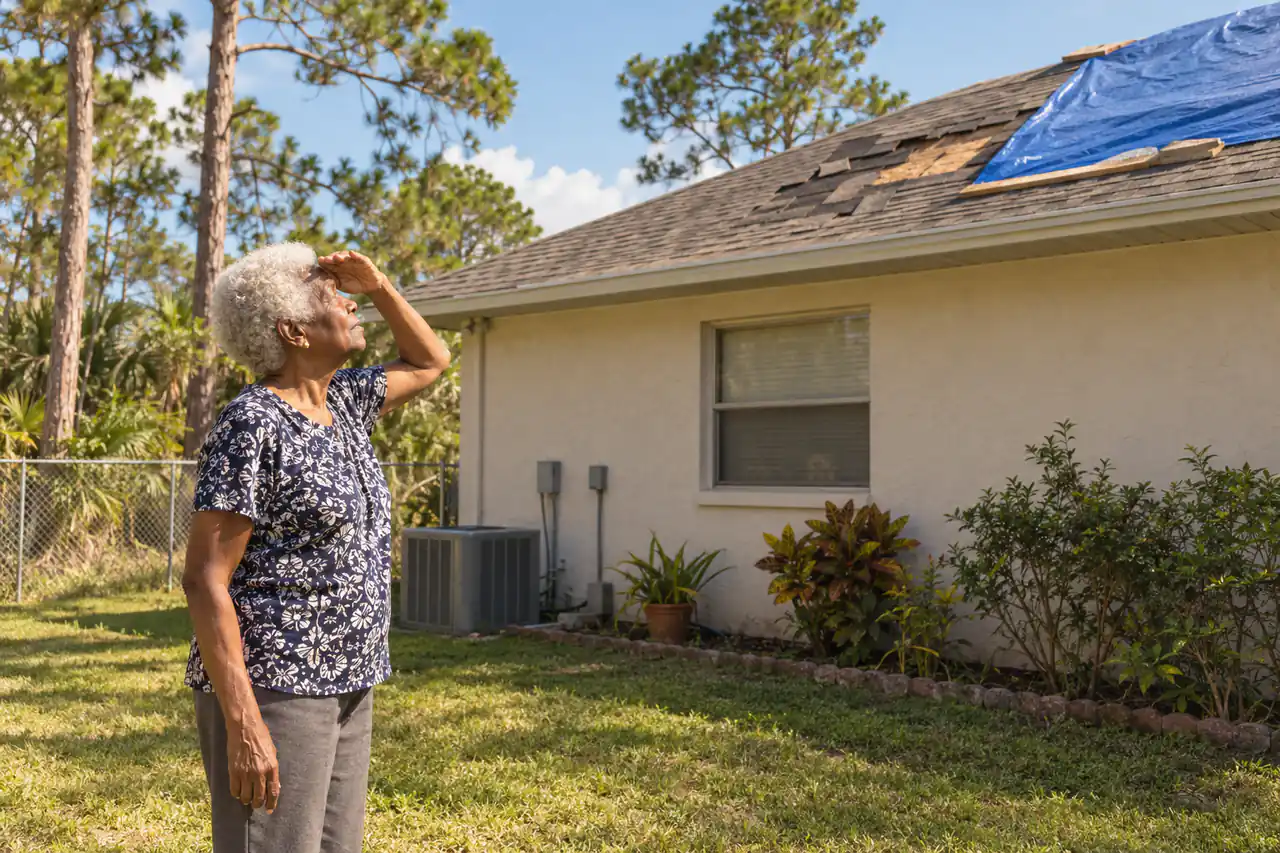 Illustrative scene: a Black woman in her seventies shading her eyes to look at missing shingles and a blue tarp on the roof of her 1980s stucco house in Palm Bay