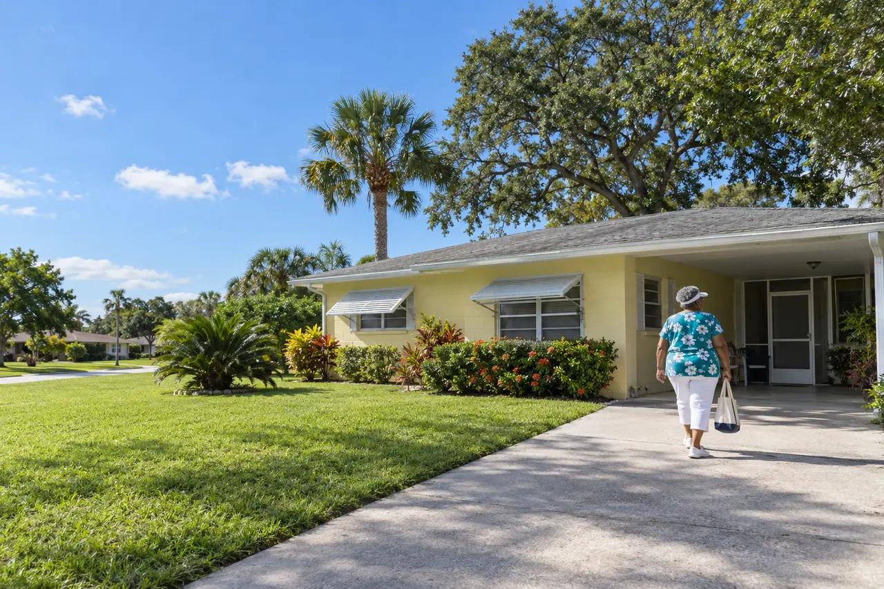 Illustrative scene: a Black woman in her seventies in a white sun visor carrying a tote bag up the driveway of her 1960s pale-yellow concrete-block ranch with a carport in Kissimmee, Florida
